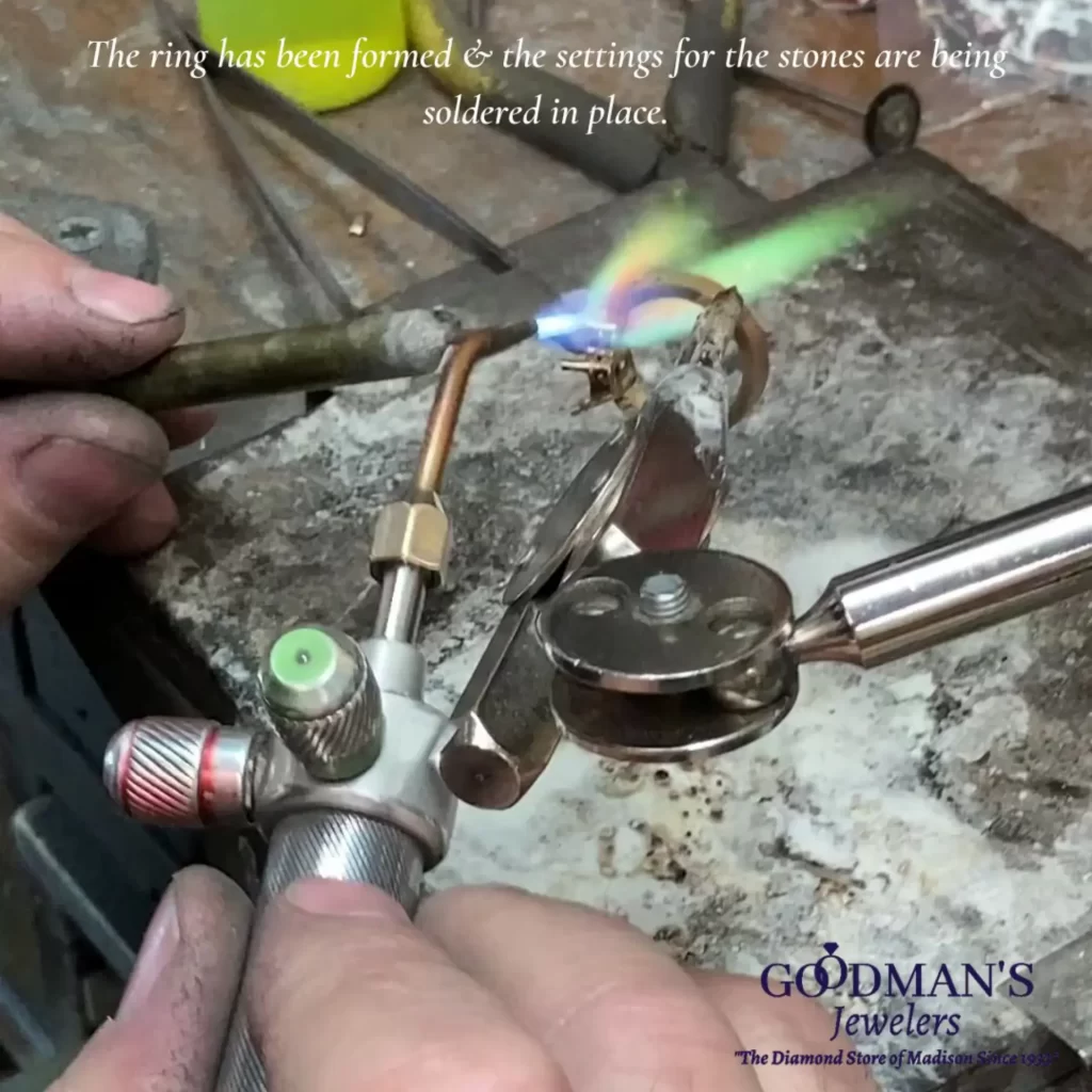 A jeweler carefully soldering the settings for gemstones onto a ring using a torch in a workshop, with tools and equipment in the background.